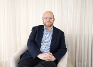 Ignia Cloud impulsa una operación de TI predictiva con servicios administrados e IA Retrato fotográfico de Esteban Rey, CEO de Ignia Cloud, sentado cómodamente en un sillón de tono claro. Viste una camisa a cuadros azules y blancos bajo un saco azul marino, lleva gafas de armazón transparente y mira de frente a la cámara con un fondo iluminado por cortinas blancas translúcidas.