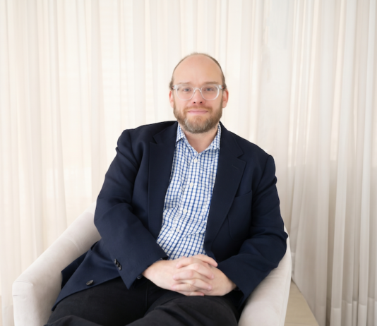Ignia Cloud impulsa una operación de TI predictiva con servicios administrados e IA Retrato fotográfico de Esteban Rey, CEO de Ignia Cloud, sentado cómodamente en un sillón de tono claro. Viste una camisa a cuadros azules y blancos bajo un saco azul marino, lleva gafas de armazón transparente y mira de frente a la cámara con un fondo iluminado por cortinas blancas translúcidas.