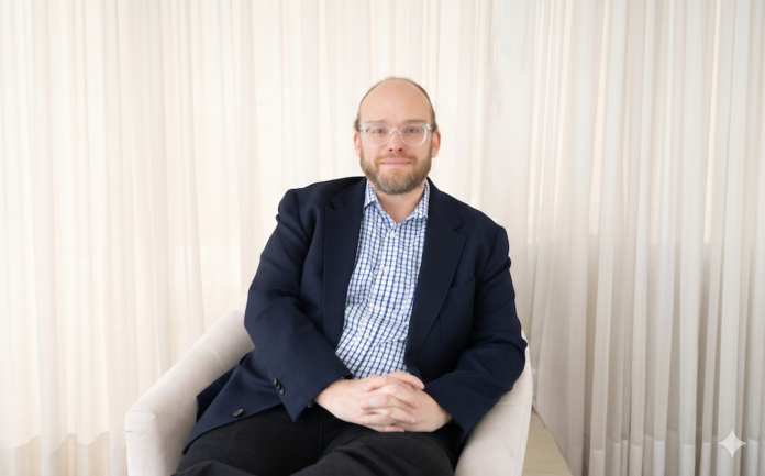 Retrato fotográfico de Esteban Rey, CEO de Ignia Cloud, sentado cómodamente en un sillón de tono claro. Viste una camisa a cuadros azules y blancos bajo un saco azul marino, lleva gafas de armazón transparente y mira de frente a la cámara con un fondo iluminado por cortinas blancas translúcidas.