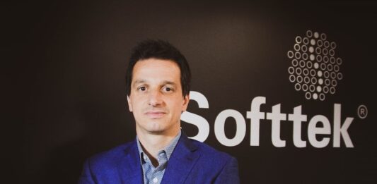 Softtek: el CIO redefine su rol y convierte la tecnología en una decisión económica Fotografía de medio cuerpo de Juan José D'Alessandro posando con los brazos cruzados. Viste una camisa de patrón claro y un saco azul, ubicado frente a un fondo oscuro donde destaca el logotipo corporativo de la empresa Softtek.
