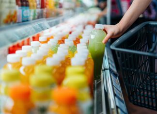 SAP destaca la sostenibilidad operativa como ventaja competitiva para las empresas Fotografía en primer plano de la mano de un comprador tomando una botella de jugo verde de una repisa refrigerada en un supermercado para colocarla dentro de una canasta de compras negra.