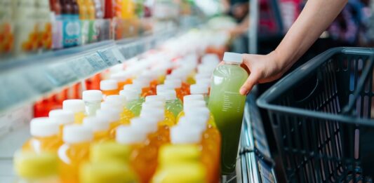 SAP destaca la sostenibilidad operativa como ventaja competitiva para las empresas Fotografía en primer plano de la mano de un comprador tomando una botella de jugo verde de una repisa refrigerada en un supermercado para colocarla dentro de una canasta de compras negra.