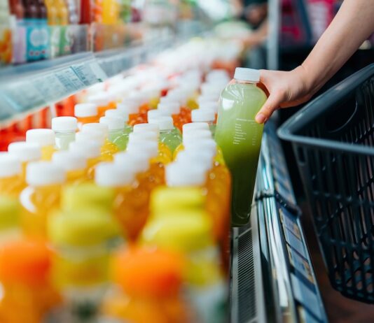 SAP destaca la sostenibilidad operativa como ventaja competitiva para las empresas Fotografía en primer plano de la mano de un comprador tomando una botella de jugo verde de una repisa refrigerada en un supermercado para colocarla dentro de una canasta de compras negra.
