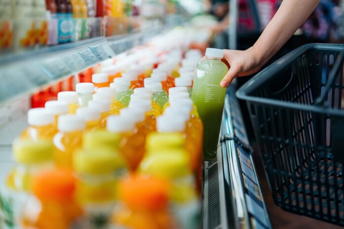Fotografía en primer plano de la mano de un comprador tomando una botella de jugo verde de una repisa refrigerada en un supermercado para colocarla dentro de una canasta de compras negra.