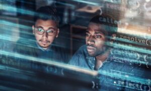 Dos especialistas en tecnología observan atentamente una pantalla en un entorno con poca luz, con proyecciones de código de programación, texto digital y flujos de datos superpuestos sobre sus rostros y el espacio de trabajo.