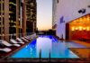 Eaton advierte sobre el reto energético para la expansión hotelera en México Fotografía de una piscina rectangular iluminada en la terraza de un edificio urbano durante el atardecer. A un costado del agua se alinea una fila de camastros blancos, mientras que el área está rodeada por altos rascacielos con ventanas iluminadas y paredes de mármol con luces cálidas.