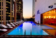 Eaton advierte sobre el reto energético para la expansión hotelera en México Fotografía de una piscina rectangular iluminada en la terraza de un edificio urbano durante el atardecer. A un costado del agua se alinea una fila de camastros blancos, mientras que el área está rodeada por altos rascacielos con ventanas iluminadas y paredes de mármol con luces cálidas.