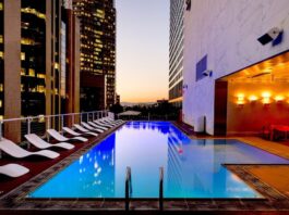 Eaton advierte sobre el reto energético para la expansión hotelera en México Fotografía de una piscina rectangular iluminada en la terraza de un edificio urbano durante el atardecer. A un costado del agua se alinea una fila de camastros blancos, mientras que el área está rodeada por altos rascacielos con ventanas iluminadas y paredes de mármol con luces cálidas.