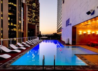 Eaton advierte sobre el reto energético para la expansión hotelera en México Fotografía de una piscina rectangular iluminada en la terraza de un edificio urbano durante el atardecer. A un costado del agua se alinea una fila de camastros blancos, mientras que el área está rodeada por altos rascacielos con ventanas iluminadas y paredes de mármol con luces cálidas.