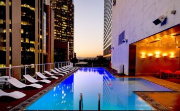Eaton advierte sobre el reto energético para la expansión hotelera en México Fotografía de una piscina rectangular iluminada en la terraza de un edificio urbano durante el atardecer. A un costado del agua se alinea una fila de camastros blancos, mientras que el área está rodeada por altos rascacielos con ventanas iluminadas y paredes de mármol con luces cálidas.