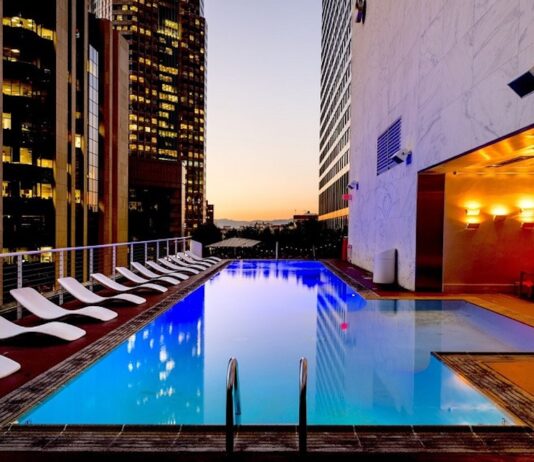 Eaton advierte sobre el reto energético para la expansión hotelera en México Fotografía de una piscina rectangular iluminada en la terraza de un edificio urbano durante el atardecer. A un costado del agua se alinea una fila de camastros blancos, mientras que el área está rodeada por altos rascacielos con ventanas iluminadas y paredes de mármol con luces cálidas.