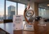 Epson México reafirma su compromiso con la sostenibilidad con el Distintivo ESR 2026 Fotografía de un premio metálico con el logotipo ESR y la inscripción CEMEFI sobre una mesa de juntas de madera. En la base del galardón se lee "EPSON MÉXICO RECEPTOR DESTACADO DEL DISTINTIVO ESR 2026". A su lado hay un folleto con el título "Epson México: Liderando con sostenibilidad" y un bloque de cristal decorativo. Al fondo, a través del amplio ventanal de la oficina, se aprecia la silueta del edificio del World Trade Center en la Ciudad de México durante el atardecer.