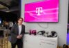 T-Systems acelera los servicios gestionados de TI con SAP Fotografía de un ejecutivo vistiendo un saco gris y camisa blanca, posando de pie en un evento corporativo o piso de exhibición. A su lado se encuentra un mueble blanco con cámaras, artículos promocionales y los logotipos de T-Systems y Google Cloud, mientras que en la pared superior destaca una pantalla proyectando el característico logotipo magenta de la marca T.