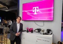 T-Systems acelera los servicios gestionados de TI con SAP Fotografía de un ejecutivo vistiendo un saco gris y camisa blanca, posando de pie en un evento corporativo o piso de exhibición. A su lado se encuentra un mueble blanco con cámaras, artículos promocionales y los logotipos de T-Systems y Google Cloud, mientras que en la pared superior destaca una pantalla proyectando el característico logotipo magenta de la marca T.