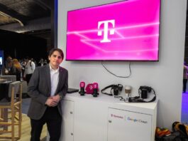 T-Systems acelera los servicios gestionados de TI con SAP Fotografía de un ejecutivo vistiendo un saco gris y camisa blanca, posando de pie en un evento corporativo o piso de exhibición. A su lado se encuentra un mueble blanco con cámaras, artículos promocionales y los logotipos de T-Systems y Google Cloud, mientras que en la pared superior destaca una pantalla proyectando el característico logotipo magenta de la marca T.