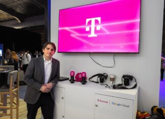 T-Systems acelera los servicios gestionados de TI con SAP Fotografía de un ejecutivo vistiendo un saco gris y camisa blanca, posando de pie en un evento corporativo o piso de exhibición. A su lado se encuentra un mueble blanco con cámaras, artículos promocionales y los logotipos de T-Systems y Google Cloud, mientras que en la pared superior destaca una pantalla proyectando el característico logotipo magenta de la marca T.