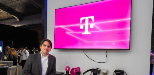 T-Systems acelera los servicios gestionados de TI con SAP Fotografía de un ejecutivo vistiendo un saco gris y camisa blanca, posando de pie en un evento corporativo o piso de exhibición. A su lado se encuentra un mueble blanco con cámaras, artículos promocionales y los logotipos de T-Systems y Google Cloud, mientras que en la pared superior destaca una pantalla proyectando el característico logotipo magenta de la marca T.