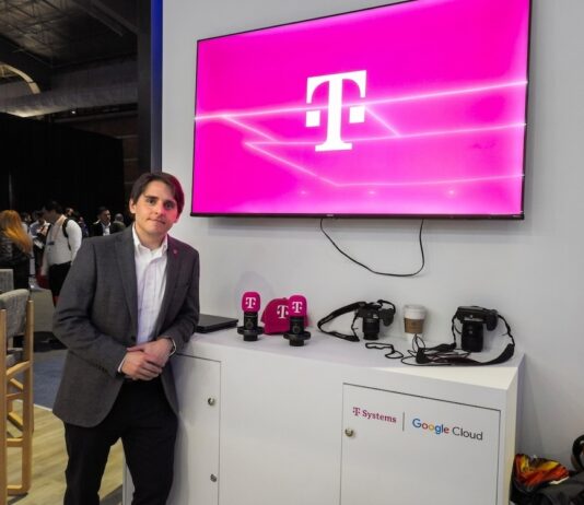 T-Systems acelera los servicios gestionados de TI con SAP Fotografía de un ejecutivo vistiendo un saco gris y camisa blanca, posando de pie en un evento corporativo o piso de exhibición. A su lado se encuentra un mueble blanco con cámaras, artículos promocionales y los logotipos de T-Systems y Google Cloud, mientras que en la pared superior destaca una pantalla proyectando el característico logotipo magenta de la marca T.