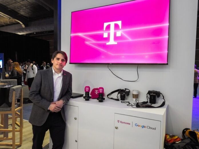 Fotografía de un ejecutivo vistiendo un saco gris y camisa blanca, posando de pie en un evento corporativo o piso de exhibición. A su lado se encuentra un mueble blanco con cámaras, artículos promocionales y los logotipos de T-Systems y Google Cloud, mientras que en la pared superior destaca una pantalla proyectando el característico logotipo magenta de la marca T.