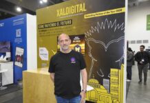 XalDigital apuesta por la transformación digital enterprise Fotografía de un directivo posando frente al stand corporativo de XalDigital, donde se exhibe su insignia como AWS Premier Partner. El módulo, diseñado en colores amarillo y negro, destaca los servicios de la compañía e incluye el mensaje "Construyendo el futuro", acompañado de gráficos urbanos y la mascota de la empresa.