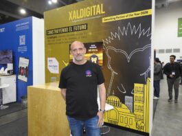 XalDigital apuesta por la transformación digital enterprise Fotografía de un directivo posando frente al stand corporativo de XalDigital, donde se exhibe su insignia como AWS Premier Partner. El módulo, diseñado en colores amarillo y negro, destaca los servicios de la compañía e incluye el mensaje "Construyendo el futuro", acompañado de gráficos urbanos y la mascota de la empresa.