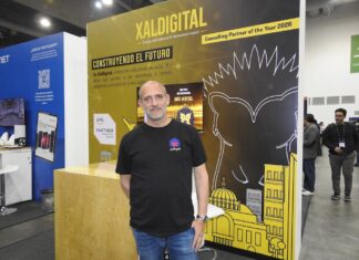 XalDigital apuesta por la transformación digital enterprise Fotografía de un directivo posando frente al stand corporativo de XalDigital, donde se exhibe su insignia como AWS Premier Partner. El módulo, diseñado en colores amarillo y negro, destaca los servicios de la compañía e incluye el mensaje "Construyendo el futuro", acompañado de gráficos urbanos y la mascota de la empresa.