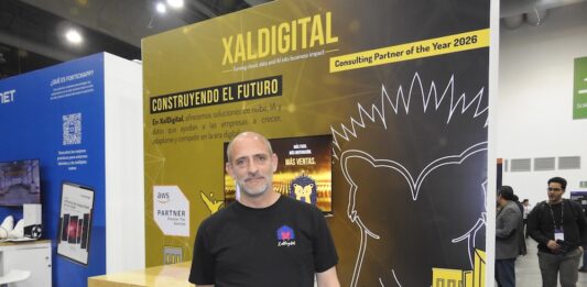 XalDigital apuesta por la transformación digital enterprise Fotografía de un directivo posando frente al stand corporativo de XalDigital, donde se exhibe su insignia como AWS Premier Partner. El módulo, diseñado en colores amarillo y negro, destaca los servicios de la compañía e incluye el mensaje "Construyendo el futuro", acompañado de gráficos urbanos y la mascota de la empresa.
