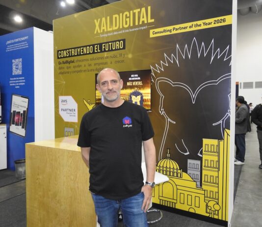 XalDigital apuesta por la transformación digital enterprise Fotografía de un directivo posando frente al stand corporativo de XalDigital, donde se exhibe su insignia como AWS Premier Partner. El módulo, diseñado en colores amarillo y negro, destaca los servicios de la compañía e incluye el mensaje "Construyendo el futuro", acompañado de gráficos urbanos y la mascota de la empresa.