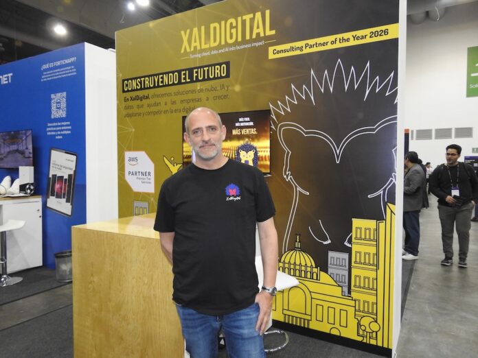 XalDigital. Arquitecturas tecnológicas alineadas a los objetivos del negocio Fotografía de un directivo posando frente al stand corporativo de XalDigital, donde se exhibe su insignia como AWS Premier Partner. El módulo, diseñado en colores amarillo y negro, destaca los servicios de la compañía e incluye el mensaje