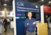 Qlik lleva la integración de datos con inteligencia artificial al siguiente nivel empresarial Fotografía de retrato de Roberto Ramírez posando con los brazos cruzados frente a un stand corporativo. Viste una camisa azul oscuro de manga larga con el logotipo de Qlik bordado. A su espalda, el panel del módulo de exhibición es de color azul y muestra en letras grandes el mensaje "Qlik. Creamos soluciones para que tus datos funcionen con AWS", acompañado del distintivo de AWS Partner. De fondo, se observa el pasillo de un centro de convenciones con múltiples asistentes y parte del stand contiguo de la marca Fastly.