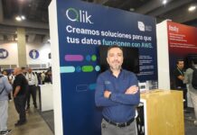 Qlik lleva la integración de datos con inteligencia artificial al siguiente nivel empresarial Fotografía de retrato de Roberto Ramírez posando con los brazos cruzados frente a un stand corporativo. Viste una camisa azul oscuro de manga larga con el logotipo de Qlik bordado. A su espalda, el panel del módulo de exhibición es de color azul y muestra en letras grandes el mensaje "Qlik. Creamos soluciones para que tus datos funcionen con AWS", acompañado del distintivo de AWS Partner. De fondo, se observa el pasillo de un centro de convenciones con múltiples asistentes y parte del stand contiguo de la marca Fastly.
