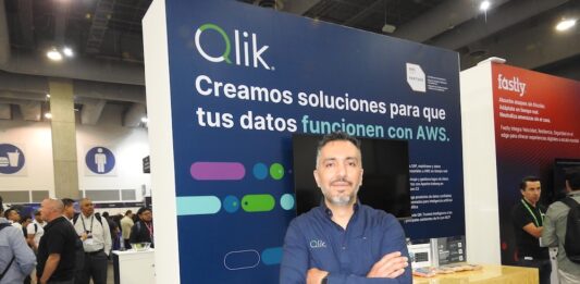 Qlik lleva la integración de datos con inteligencia artificial al siguiente nivel empresarial Fotografía de retrato de Roberto Ramírez posando con los brazos cruzados frente a un stand corporativo. Viste una camisa azul oscuro de manga larga con el logotipo de Qlik bordado. A su espalda, el panel del módulo de exhibición es de color azul y muestra en letras grandes el mensaje "Qlik. Creamos soluciones para que tus datos funcionen con AWS", acompañado del distintivo de AWS Partner. De fondo, se observa el pasillo de un centro de convenciones con múltiples asistentes y parte del stand contiguo de la marca Fastly.
