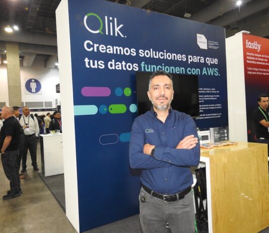Qlik lleva la integración de datos con inteligencia artificial al siguiente nivel empresarial Fotografía de retrato de Roberto Ramírez posando con los brazos cruzados frente a un stand corporativo. Viste una camisa azul oscuro de manga larga con el logotipo de Qlik bordado. A su espalda, el panel del módulo de exhibición es de color azul y muestra en letras grandes el mensaje "Qlik. Creamos soluciones para que tus datos funcionen con AWS", acompañado del distintivo de AWS Partner. De fondo, se observa el pasillo de un centro de convenciones con múltiples asistentes y parte del stand contiguo de la marca Fastly.