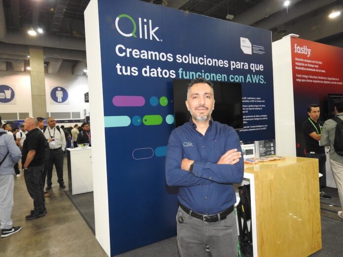 Fotografía de retrato de Roberto Ramírez posando con los brazos cruzados frente a un stand corporativo. Viste una camisa azul oscuro de manga larga con el logotipo de Qlik bordado. A su espalda, el panel del módulo de exhibición es de color azul y muestra en letras grandes el mensaje 