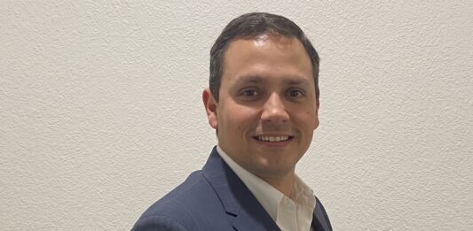 Tanium: Agentes de IA; ¿Por qué la autonomía sin datos fiables sólo genera errores a gran escala? Fotografía de retrato de Heriberto Cabrera mirando hacia la cámara y sonriendo levemente. Viste un saco de vestir azul marino sobre una camisa blanca de cuello abierto, posando de perfil tres cuartos frente a una pared texturizada de color blanco.