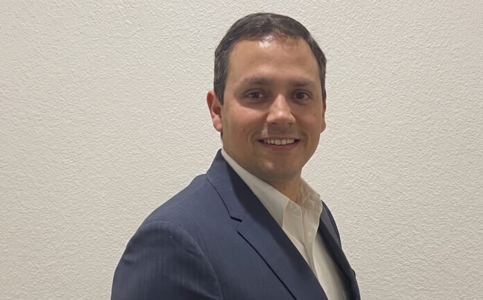Heriberto Cabrera aborda la transición hacia la IA de ejecución en ciberseguridad Fotografía de retrato de Heriberto Cabrera mirando hacia la cámara y sonriendo levemente. Viste un saco de vestir azul marino sobre una camisa blanca de cuello abierto, posando de perfil tres cuartos frente a una pared texturizada de color blanco.