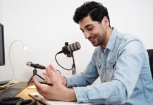 ElevenLabs utiliza IA para preservar la voz de pacientes ante enfermedades que afectan el habla Fotografía de un hombre sonriendo mientras habla frente a un micrófono de estudio montado en un brazo articulado. Viste una camisa de mezclilla azul y se encuentra sentado frente a un escritorio de madera, gesticulando de manera relajada con una mano sobre un cuaderno abierto. En la mesa también se aprecian un teclado, audífonos y el borde de un monitor.