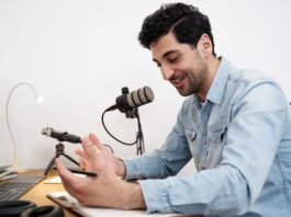 ElevenLabs utiliza IA para preservar la voz de pacientes ante enfermedades que afectan el habla Fotografía de un hombre sonriendo mientras habla frente a un micrófono de estudio montado en un brazo articulado. Viste una camisa de mezclilla azul y se encuentra sentado frente a un escritorio de madera, gesticulando de manera relajada con una mano sobre un cuaderno abierto. En la mesa también se aprecian un teclado, audífonos y el borde de un monitor.