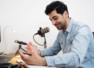 ElevenLabs utiliza IA para preservar la voz de pacientes ante enfermedades que afectan el habla Fotografía de un hombre sonriendo mientras habla frente a un micrófono de estudio montado en un brazo articulado. Viste una camisa de mezclilla azul y se encuentra sentado frente a un escritorio de madera, gesticulando de manera relajada con una mano sobre un cuaderno abierto. En la mesa también se aprecian un teclado, audífonos y el borde de un monitor.