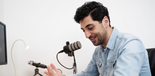 ElevenLabs utiliza IA para preservar la voz de pacientes ante enfermedades que afectan el habla Fotografía de un hombre sonriendo mientras habla frente a un micrófono de estudio montado en un brazo articulado. Viste una camisa de mezclilla azul y se encuentra sentado frente a un escritorio de madera, gesticulando de manera relajada con una mano sobre un cuaderno abierto. En la mesa también se aprecian un teclado, audífonos y el borde de un monitor.