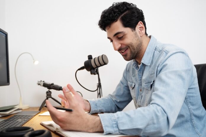 Fotografía de un hombre sonriendo mientras habla frente a un micrófono de estudio montado en un brazo articulado. Viste una camisa de mezclilla azul y se encuentra sentado frente a un escritorio de madera, gesticulando de manera relajada con una mano sobre un cuaderno abierto. En la mesa también se aprecian un teclado, audífonos y el borde de un monitor.