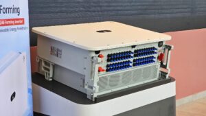 Fotografía en primer plano de un inversor solar comercial Huawei SUN2000 colocado sobre una base blanca. El equipo metálico muestra múltiples conectores fotovoltaicos en su panel frontal con tapas azules y rojas, ventilaciones inferiores y etiquetas de seguridad en el chasis.