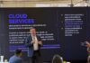 KIO IT Services revela las lecciones tecnológicas que dejó el Mundial 2026 Fotografía de Bruno Juanes hablando por micrófono frente a una pantalla oscura de presentación que resalta el texto "CLOUD SERVICES" y sus beneficios operativos, mientras miembros de la audiencia lo escuchan y graban con sus teléfonos móviles.