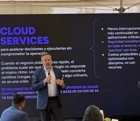 KIO IT Services revela las lecciones tecnológicas que dejó el Mundial 2026 Fotografía de Bruno Juanes hablando por micrófono frente a una pantalla oscura de presentación que resalta el texto "CLOUD SERVICES" y sus beneficios operativos, mientras miembros de la audiencia lo escuchan y graban con sus teléfonos móviles.