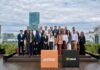 Inter desembarca en la Argentina con una plataforma integral para inversiones internacionales Fotografía de grupo al aire libre en una terraza donde ejecutivos y ejecutivas posan sonrientes organizados en dos filas. En el primer plano destacan dos tótems cúbicos: uno de color naranja con el logotipo de Inter y otro de color negro con la marca Bind, con edificios urbanos de fondo bajo un cielo nublado.