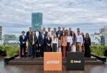 Inter desembarca en la Argentina con una plataforma integral para inversiones internacionales Fotografía de grupo al aire libre en una terraza donde ejecutivos y ejecutivas posan sonrientes organizados en dos filas. En el primer plano destacan dos tótems cúbicos: uno de color naranja con el logotipo de Inter y otro de color negro con la marca Bind, con edificios urbanos de fondo bajo un cielo nublado.