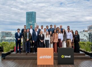 Inter desembarca en la Argentina con una plataforma integral para inversiones internacionales Fotografía de grupo al aire libre en una terraza donde ejecutivos y ejecutivas posan sonrientes organizados en dos filas. En el primer plano destacan dos tótems cúbicos: uno de color naranja con el logotipo de Inter y otro de color negro con la marca Bind, con edificios urbanos de fondo bajo un cielo nublado.