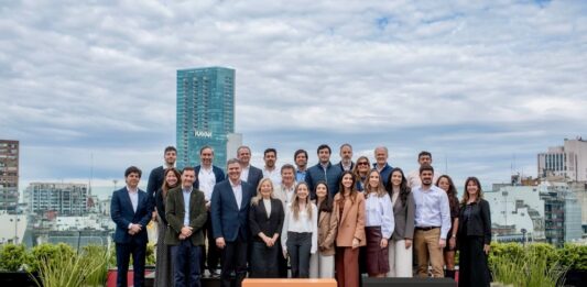 Inter desembarca en la Argentina con una plataforma integral para inversiones internacionales Fotografía de grupo al aire libre en una terraza donde ejecutivos y ejecutivas posan sonrientes organizados en dos filas. En el primer plano destacan dos tótems cúbicos: uno de color naranja con el logotipo de Inter y otro de color negro con la marca Bind, con edificios urbanos de fondo bajo un cielo nublado.