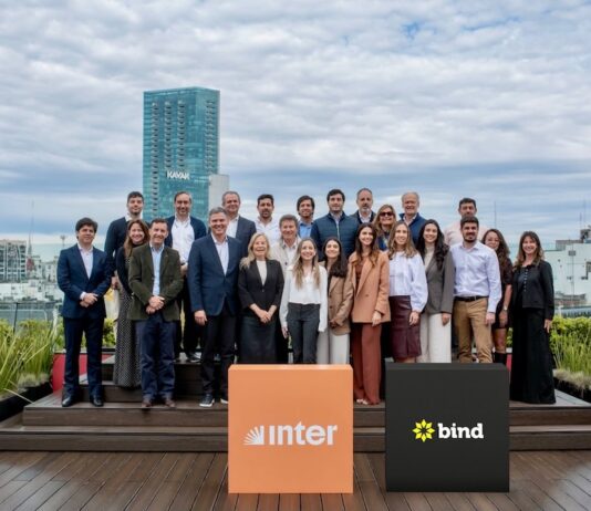 Inter desembarca en la Argentina con una plataforma integral para inversiones internacionales Fotografía de grupo al aire libre en una terraza donde ejecutivos y ejecutivas posan sonrientes organizados en dos filas. En el primer plano destacan dos tótems cúbicos: uno de color naranja con el logotipo de Inter y otro de color negro con la marca Bind, con edificios urbanos de fondo bajo un cielo nublado.