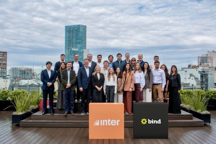 Fotografía de grupo al aire libre en una terraza donde ejecutivos y ejecutivas posan sonrientes organizados en dos filas. En el primer plano destacan dos tótems cúbicos: uno de color naranja con el logotipo de Inter y otro de color negro con la marca Bind, con edificios urbanos de fondo bajo un cielo nublado.
